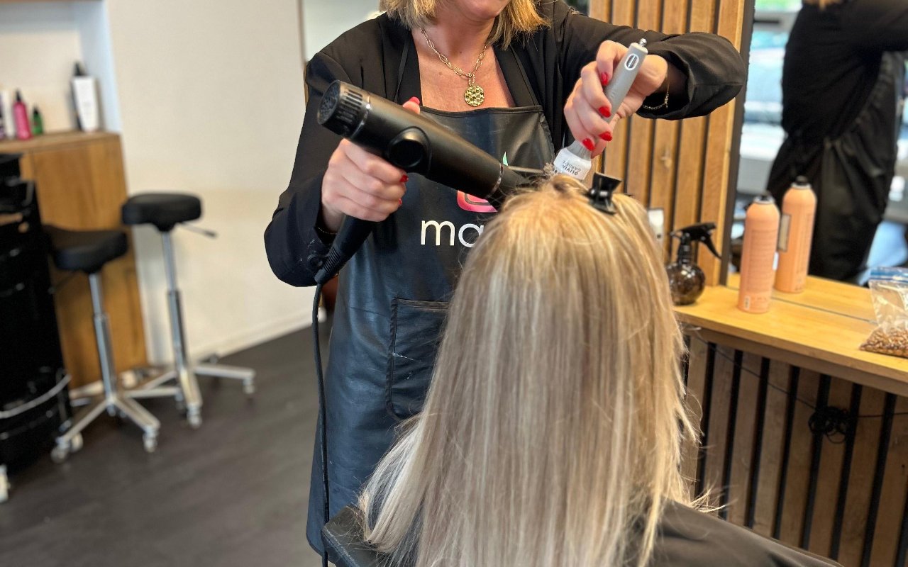Een styliste föhnt het blonde haar van een klant bij de spiegel